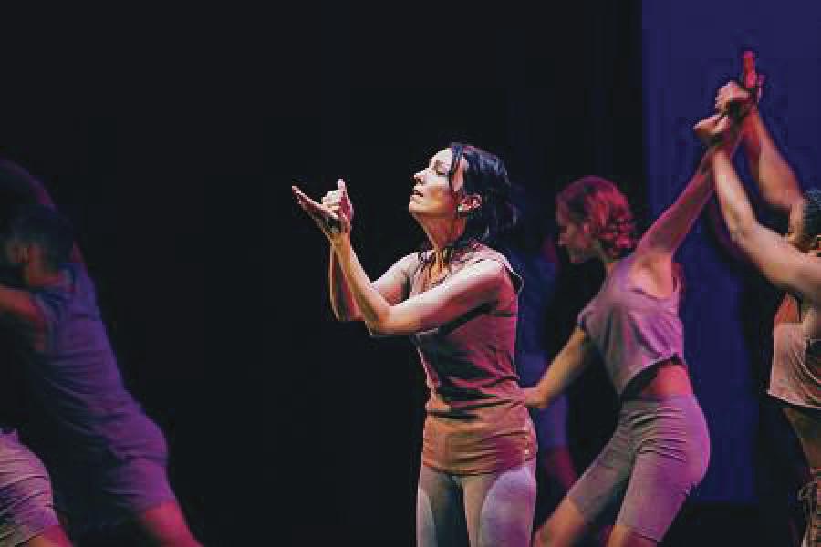 In the center, a woman stands in profile. She is highlighted by a spotlight. There are several performers on stage posing in the background.