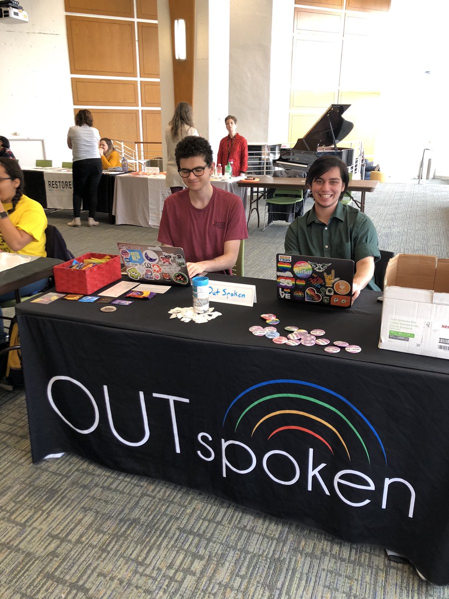 Colton and Ryan sitting at a table in the Fireside Lounge. The table is covered with a black tablecloth with the OUTspoken logo on the front, and there are pins, stickers, and contraceptives on the table.