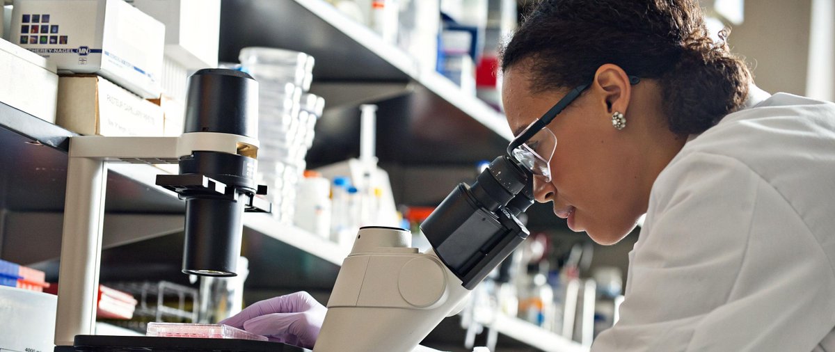 A woman in a lab coat, gloves, and goggles looking through a high powered microscope.