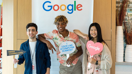 Three CSSI students posing in front of a Google sign holding props reading, "I make coding cool," "I look like a computer scientist," and "Google."