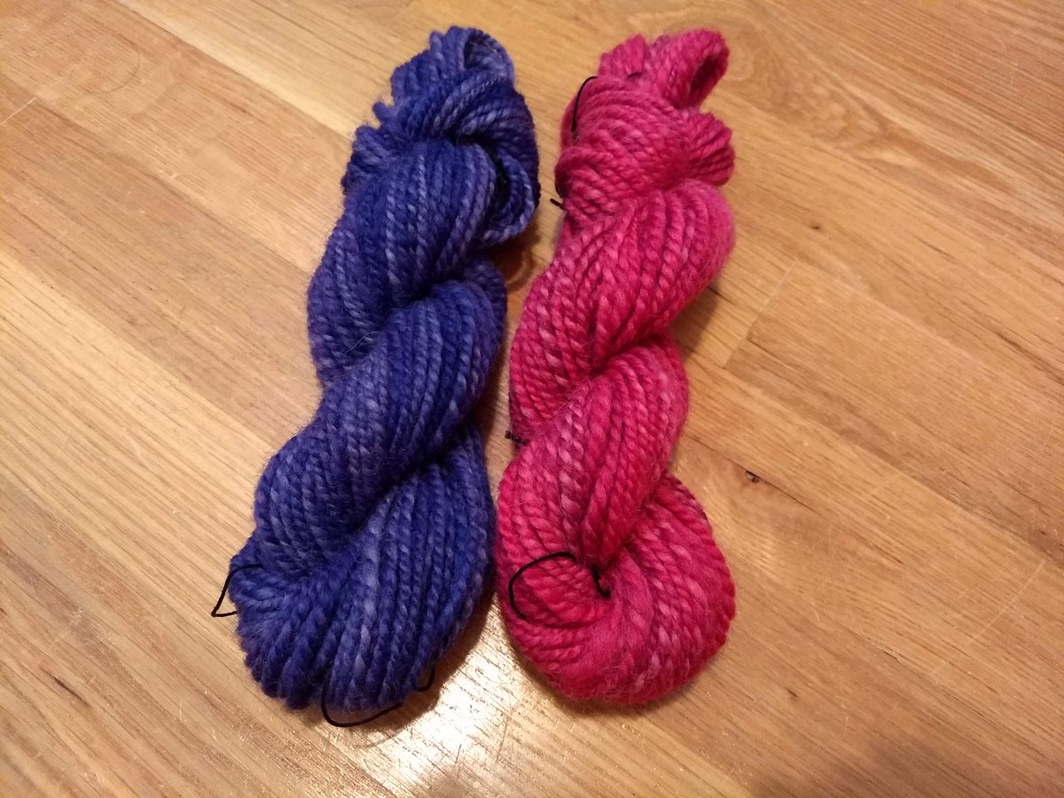 Two skeins of yarn, neatly twisted back on themselves. They are spun identically, as a consistent smooth 2-ply. One is dyed a blueish purple, the other a bright pink. Both have some paler shades mixed throughout.