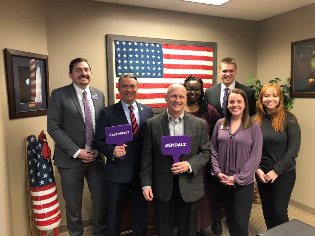 Congressman Don Bacon and five advocates stand in front of an American flag. Two advocates are holding signs. One says “#ENDALZ” and the other says “@ALZIMPACT”.