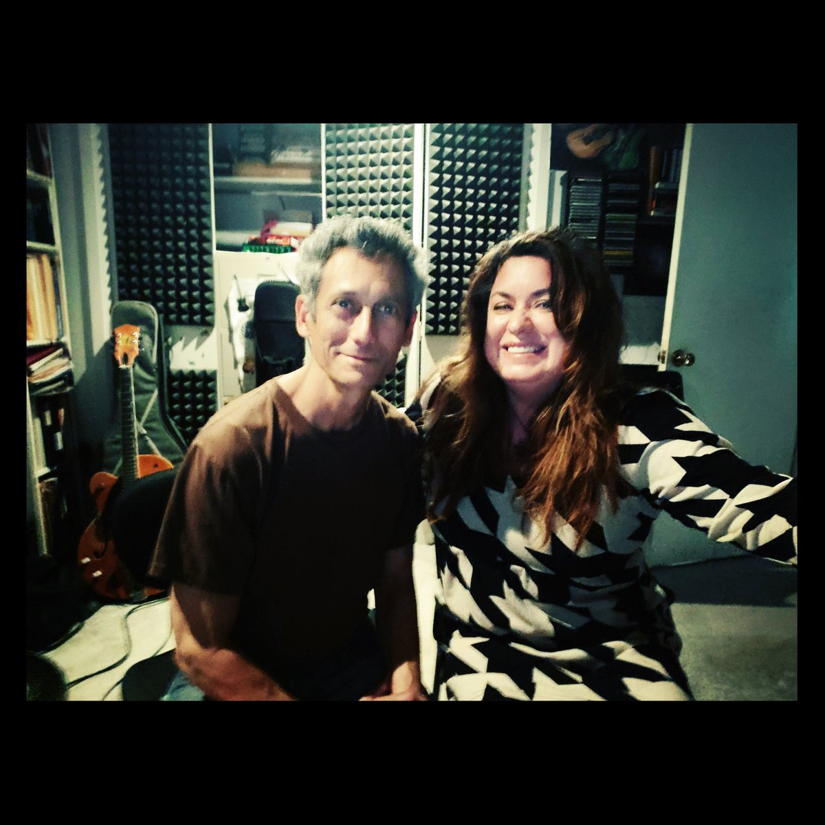 Monte and Amy taking a selfie in the recording studio. They are both smiling. Guitars are in the background. The walls have soundproofing foam on them.