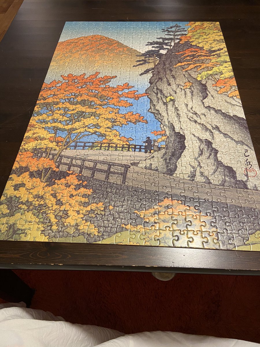 A jigsaw puzzle is on a brown wooden table. The puzzle is complete. It shows a beautiful artwork by Kawase Hasui, a woodblock print called Autumn at Saruiwa, Shiobara, 1949, a tight curve of mountain road amidst maple trees turning from green to yellow to flaming orange against a breathtakingly beautiful blue sky. Two small figures on the road admire the gorge and mountain on the far side clothed in autumn’s orange.