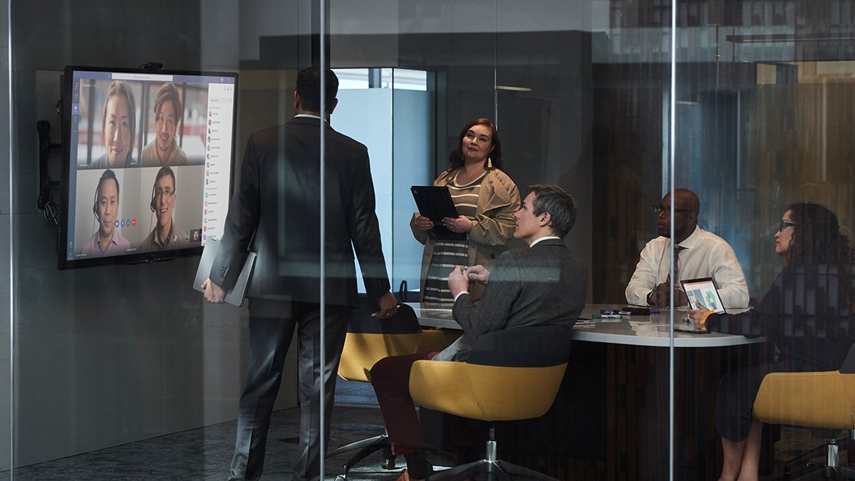 5 white collar employees are in a meeting room. They are all watching a large screen which is displaying a Microsoft Teams interface, with 4 people on it in a video call with said white caollar employees.