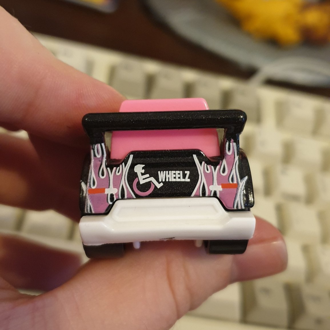 photo of the back of the same small diecast wheelchair. it has a black back cross bar, a pink and white flame design, a pink and white icon of a wheelchair user wearing a sports helmet, and the word 'wheelz'.