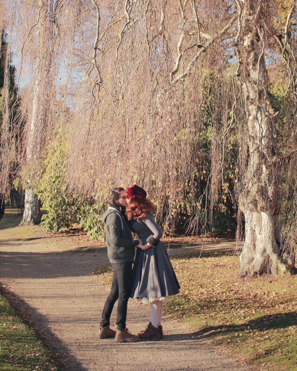 Image of Jessie and Claud holding hands and kissing in a sunny park beside some trees. Jessie is wearing a grey dress and red head wrap and Claud is in a grey jumper and black jeans.