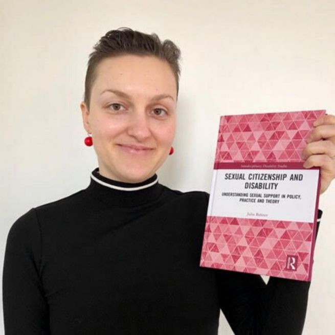 Julia is wearing a black polo neck jumper, red bauble earrings and is holding up her book which has a pink hexagon design on the cover