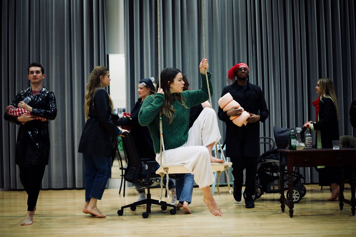 Actor sitting on swing while others walk in background in rehearsal room"