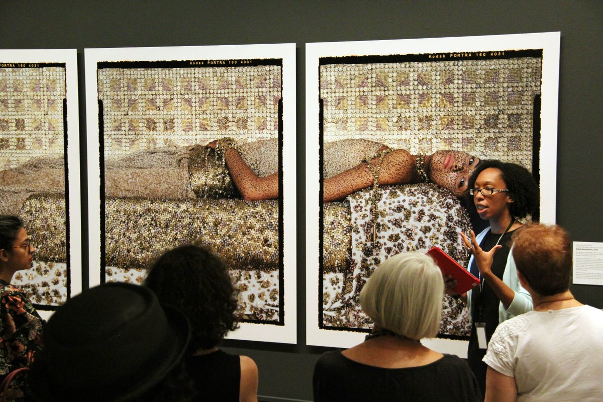 A NMWA staff member gives a gallery talk of "She Who Tells a Story" in front of a crowd. The staff member is standing beside a large triptych of a woman lying down.