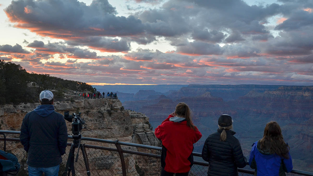 Several people wearing winter coats are viewing a colorful sunset of patchy clouds above a vast canyon landscape. NPS/M.Quinn