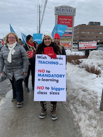 Teacher on the strike line holding a sign with the text “Wish I was teaching, no to mandatory e-learning, No to bigger class sizes.”