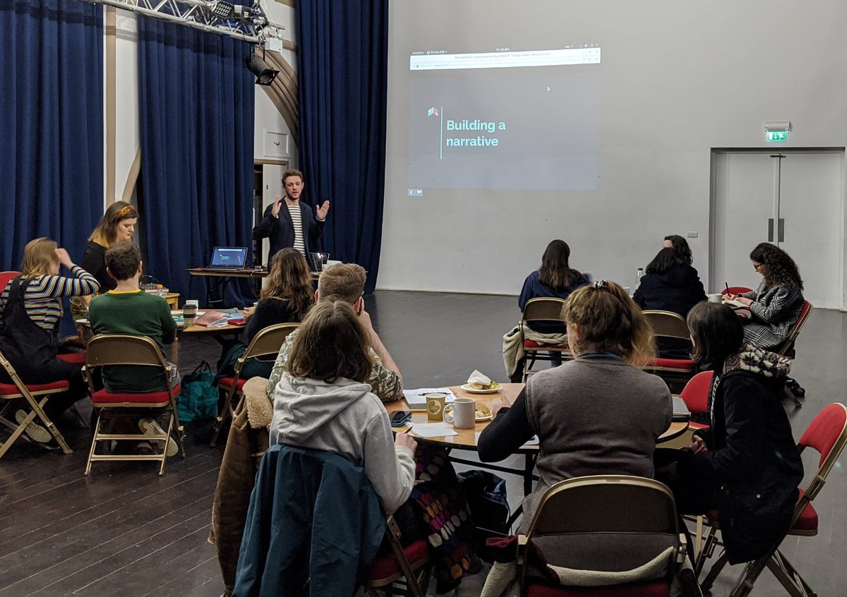 Lots of people sat around cabaret tables, facing a presentation screen with the words 'Building a narrative'