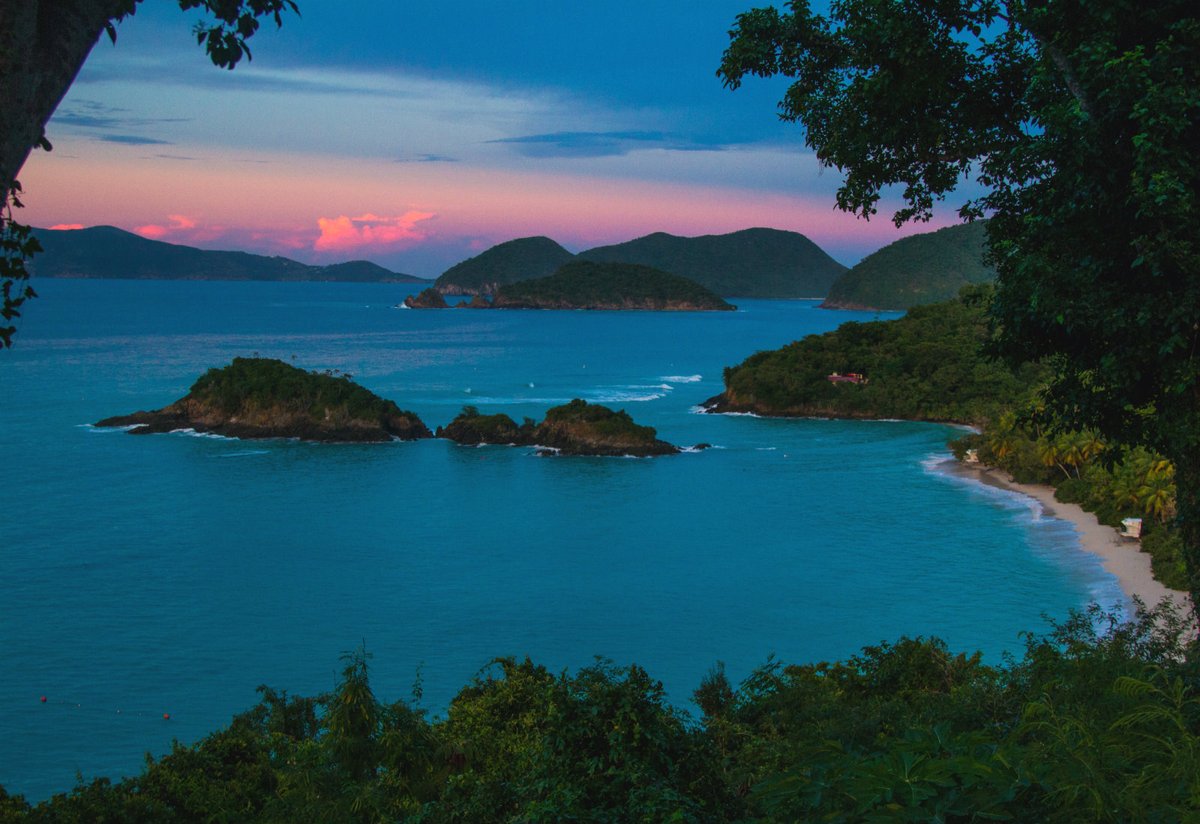 A long sandy beach curves along the edge of a jungle covered island next to blue ocean waters under a colorful sunset sky.