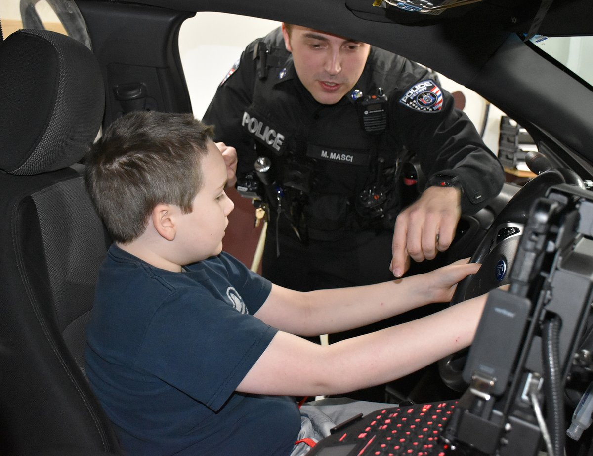 An elementary boy is sitting in the driver seat of a police car. A male police officer is pointing to something on the steering wheel. 