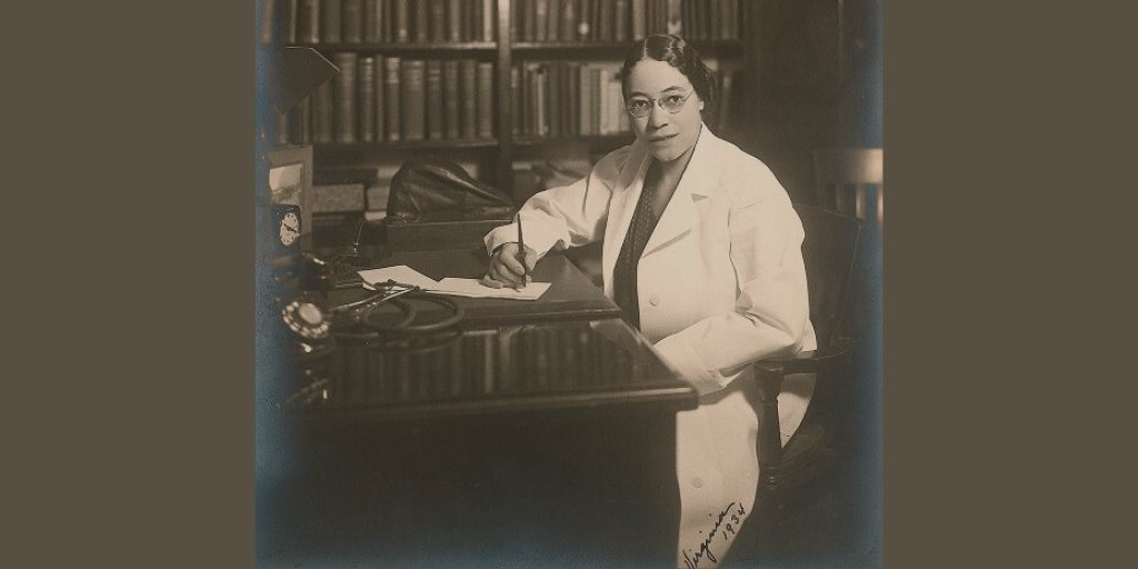 Sepia-toned photo of an African-American woman sitting at a desk. She wears a white doctor's coat. Behind her is a large, filled bookshelf.