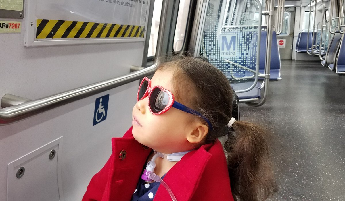 photo of young girl with trach and sunglasses sitting in wheelchair looking out the window of DC Metro train