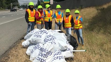 Six teens in jeans, orange safety vests with yellow reflective stripes, and green helmets give thumbs-up gestures while posing behind a knee-high pile of filled Ecology Youth Corps plastic trash bags, in a grassy area next to the shoulder of a freeway.