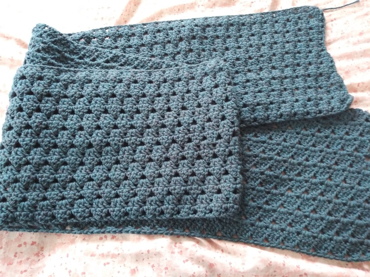 A dark teal crocheted scarf on a pastel pink background, folded in half and partly folded over again to show the entire scarf.