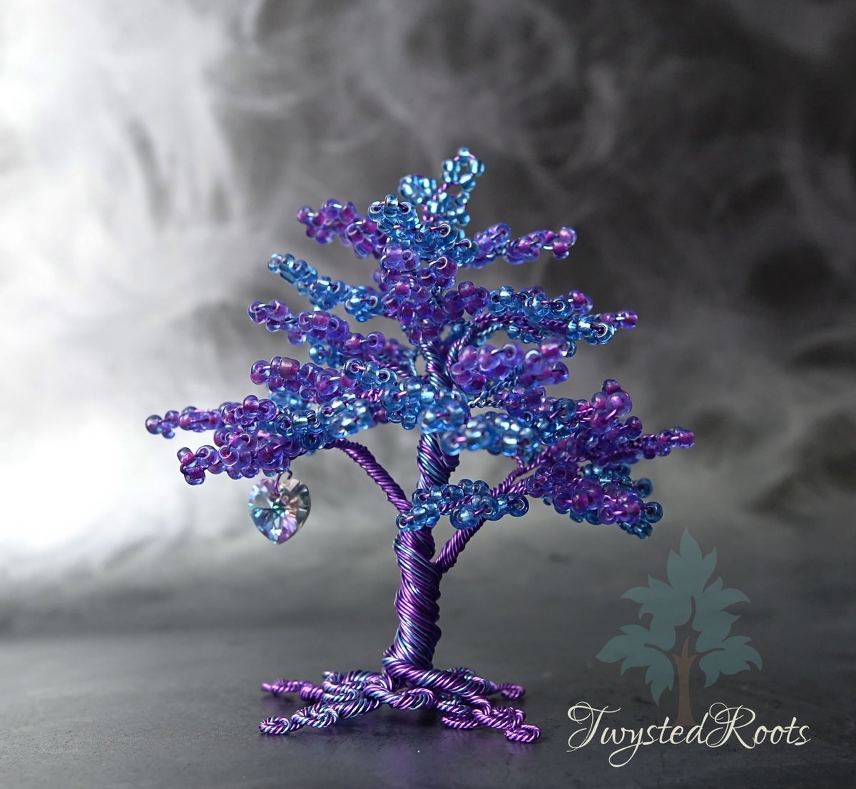 Blue and purple bead and wire tree sculpture with a violet Swarovski heart hanging from a lower left branch. Background is a misty grey effect. Twysted Roots logo is in the bottom right corner. 