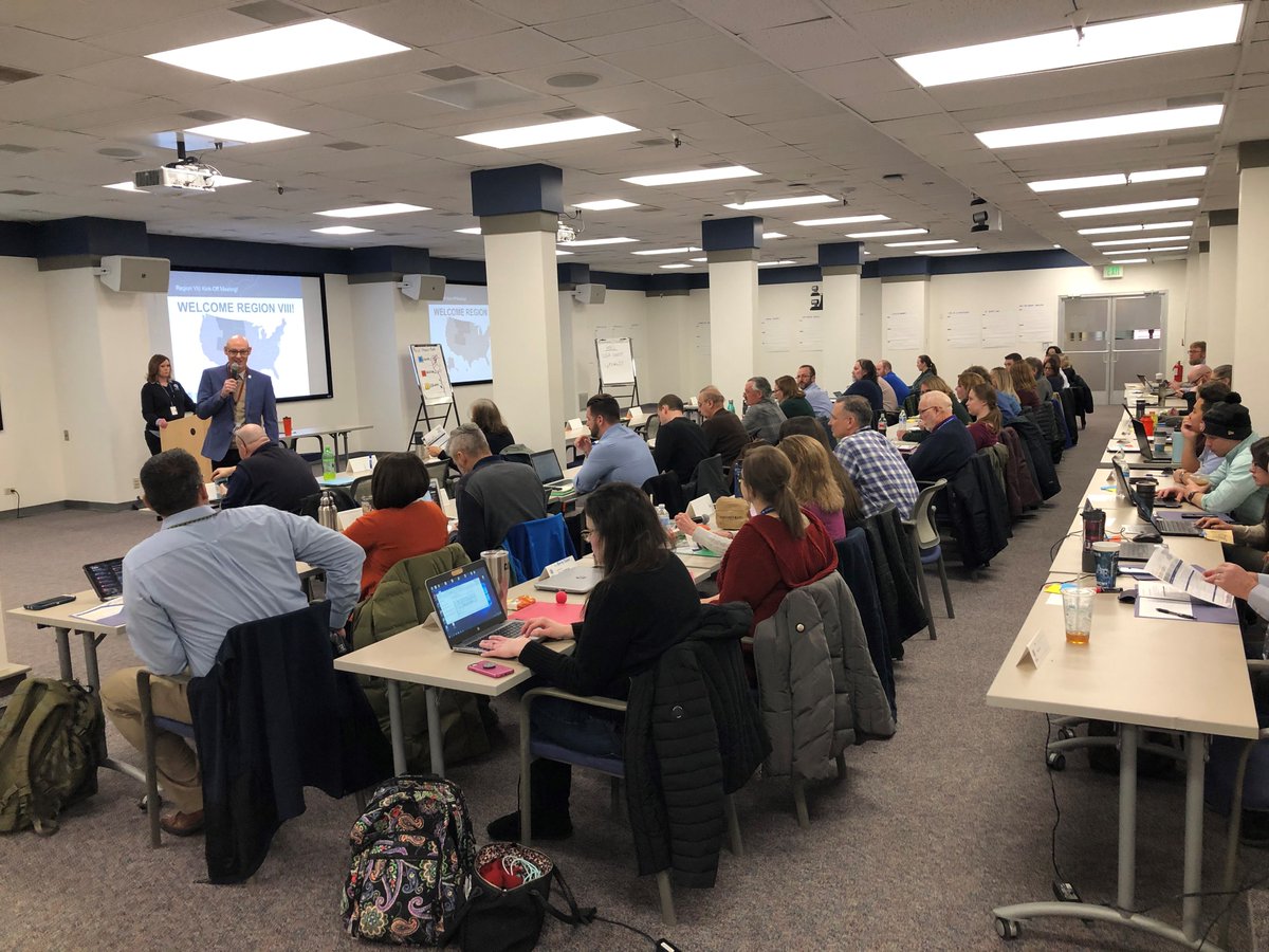 a group of people are sitting at tables listening to a man in a blue suit talking to them at the front of the room, holding a microphone.