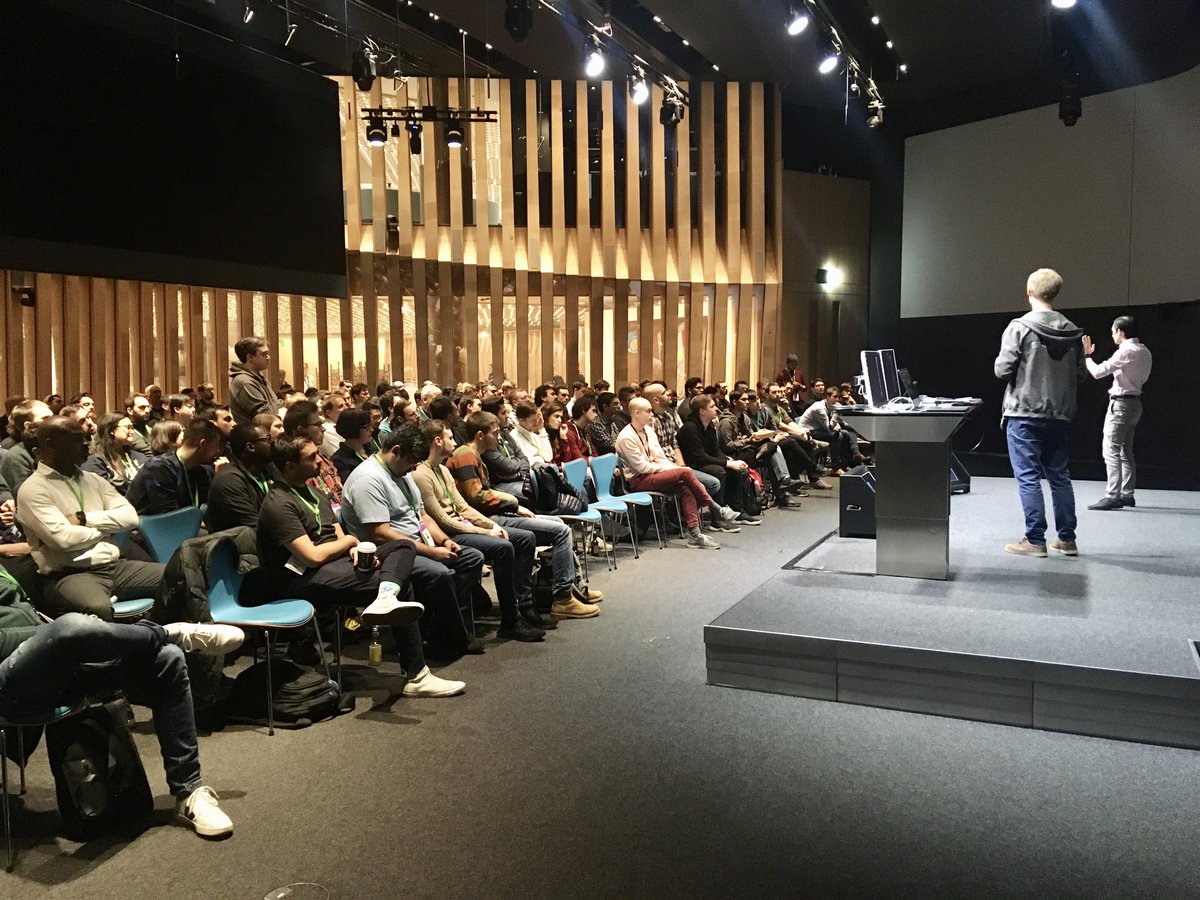 Photo of people in an auditorium taken from the side of the stage. There are two speakers on the stage.