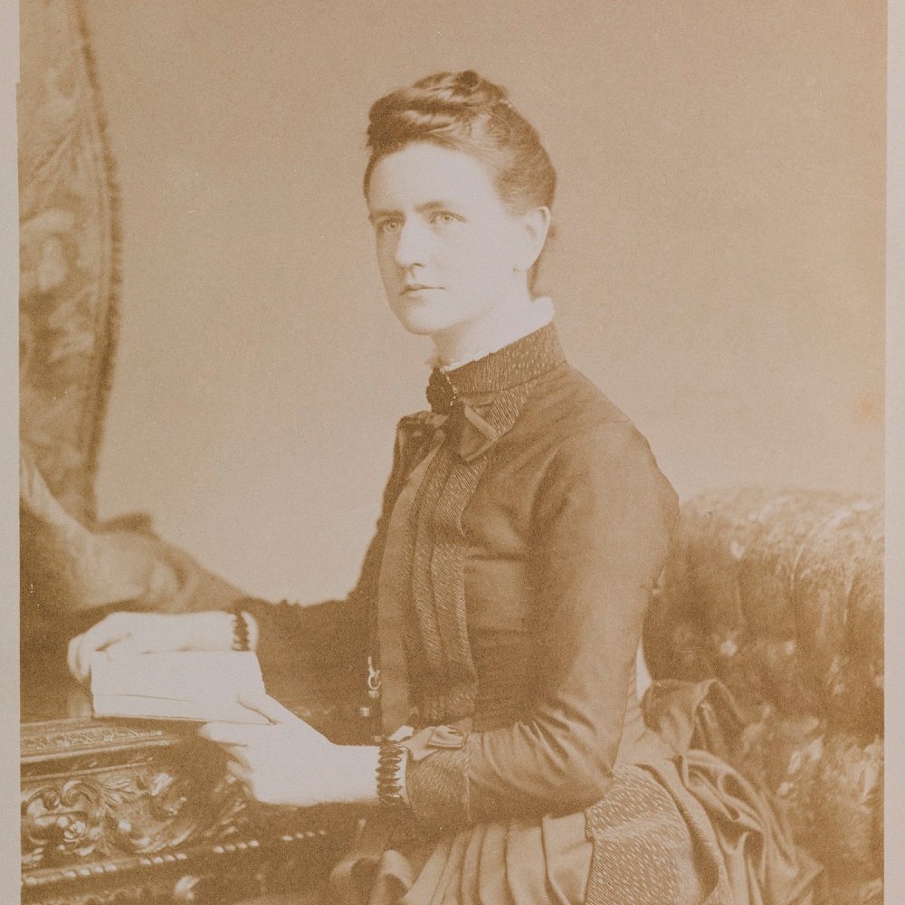 sepia photo of a young white Victorian woman in a black dress, sitting at a desk with an open book in her hands