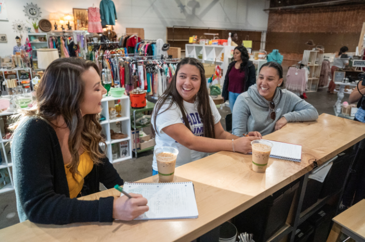 Three college-aged women are sitting at a coffee bar. They are laughing and writing in notebooks.