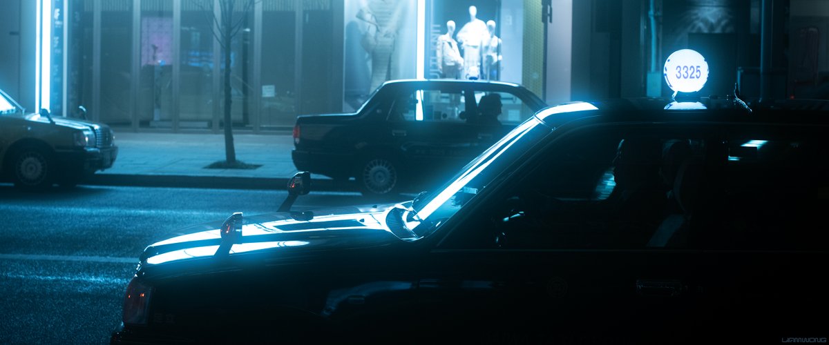Photography by Liam Wong of Tokyo at night in the Ginza district. A taxi waits at a rank. The silhouette of the drivers face can be seen. The lights from the previous building are reflecting strongly on the bonnet of the taxi. The light is piercing and sharp on the eyes. It has a blue tint.
