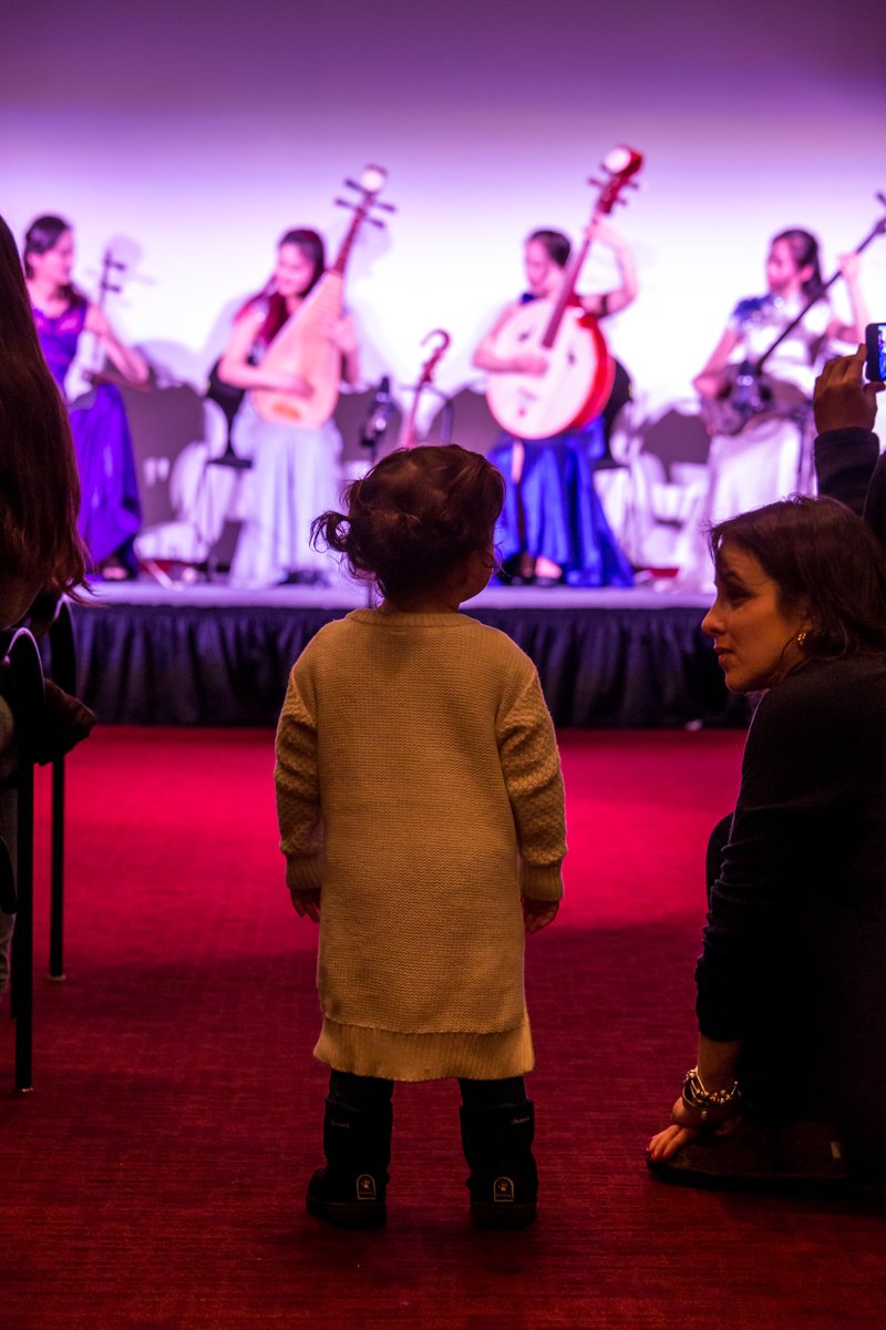 Child, seen from behind, stands in a theater looking up at a stage on which performers in long gowns or robes play stringed instruments. An adult kneels to speak to the child. 