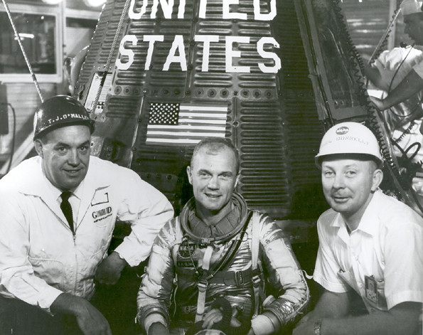 Three men pose in front of a rocket capsule with "United States" written on it.  