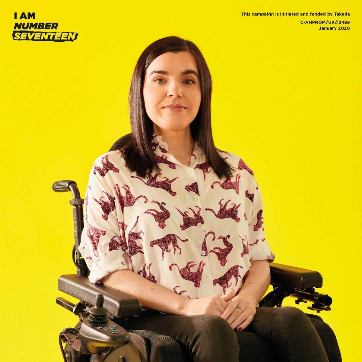 Emma is sitting in her powerchair in front of a yellow background with writing in the top left corner "I am number seventeen". Emma is wearing a white shirt with pink leopards and black skinny jeans. She has her dark brown hair down just below her collarbones. 