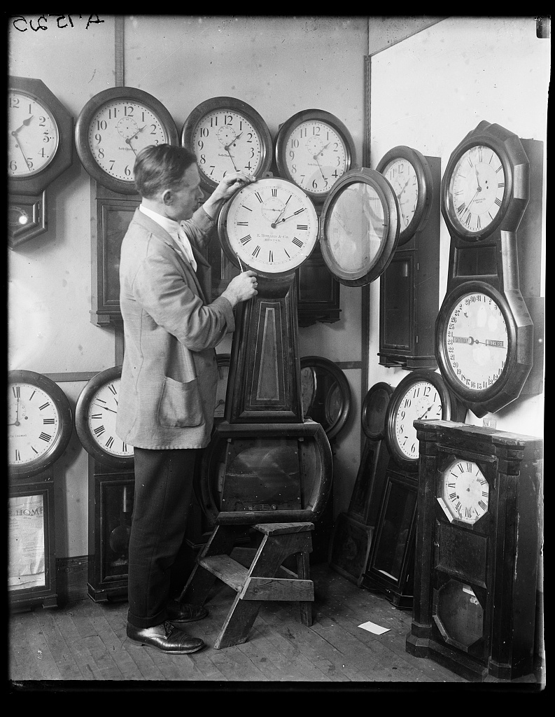A man standing on the left of a clock surrounded by a wall of clocks