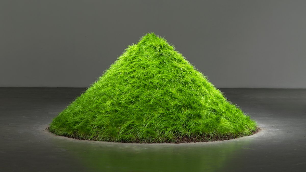 A photo of Hans Haacke's work "Grass Grows" as it appears in his exhibition "All Connected." The work is composed of a conical mound of dirt covered in a bright green carpet of grass.