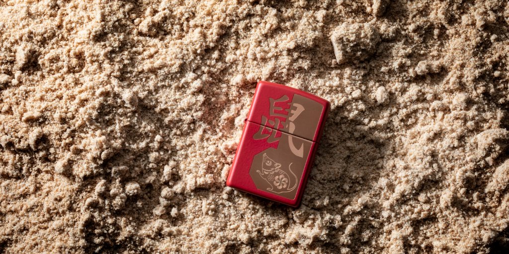 A red matte Zippo lighter decorated in celebration of the Year of the Rat lies on a sandy background.