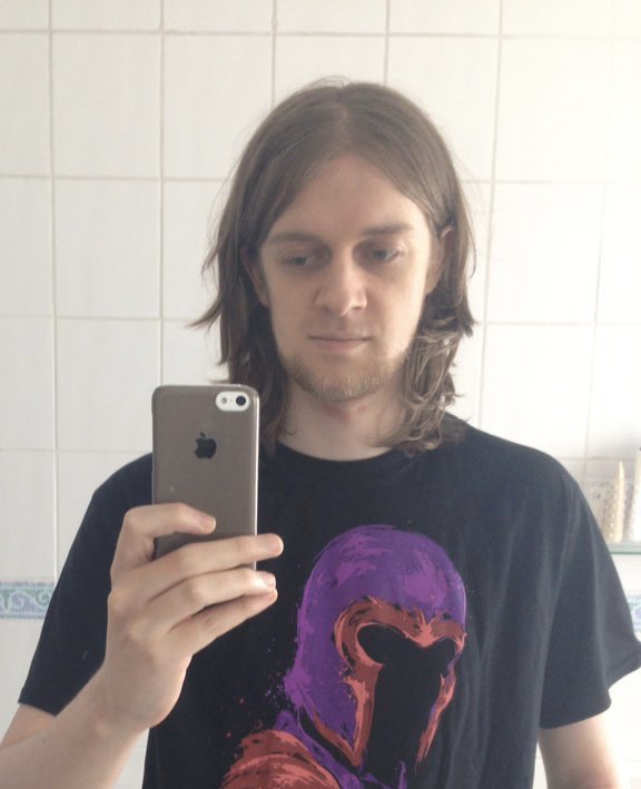 A photo of Mia pre-transition with shaggy brown hair and facial hair taking a mirror selfie in the bathroom.