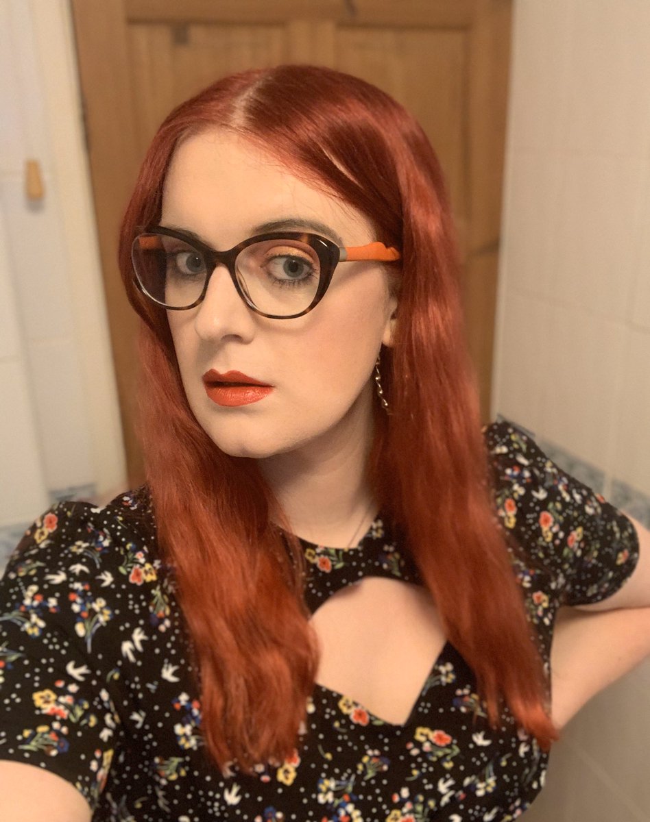 Mia as she looks now with long red hair, cat eye glasses, a black floral dress and red lipstick.