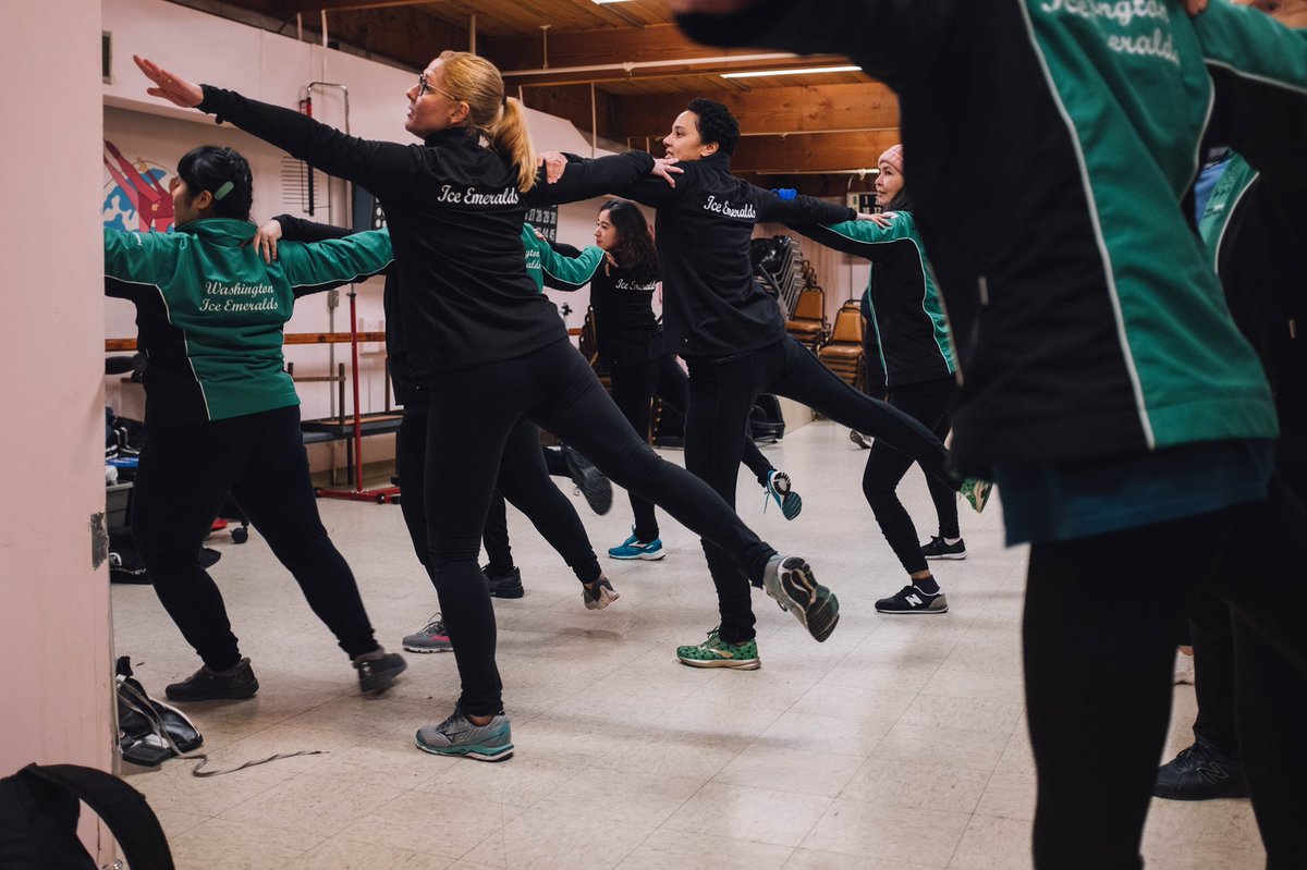 Off ice synchronized ice skating practice. People in rows holding shoulders with their right legs extended behind them.