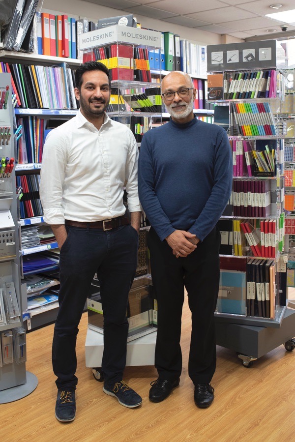 Qusai & Hafiz Jafferji standing in the shop, with shelves crammed with multicoloured array of stationery products all around them