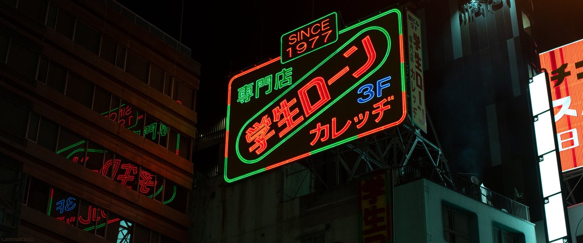Photography by Liam Wong of Tokyo at night. A large neon sign attached to a building. It says '1977' and is red, green and blue. It feels retro and the colors are desaturated to fit the vibe. Smoke from a yakitori grill restaurant is seen behind the neon.