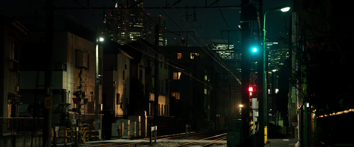 Photography by Liam Wong of Tokyo at night. An empty train track with Shinjuku in the background. It is green and yellow in color with the signal lights in red. It feels hazy.
