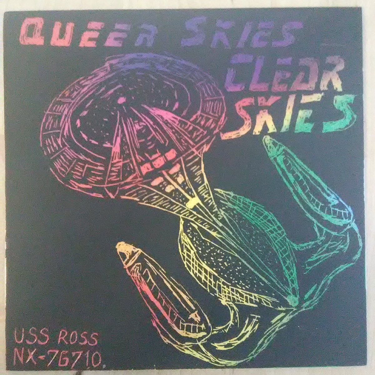 Black background rainbow gradiant colors revealed by scratches.
Across the top says "QUEER SKIES CLEAR SKIES" at the bottom says "USS ROSS NX-76710"
In the middle is a crudely sketched Star Trek style space ship seen from above and behind. Ship has a large front saucer section, swooping down to a relatively wide body with two nacelle extensions to either side.