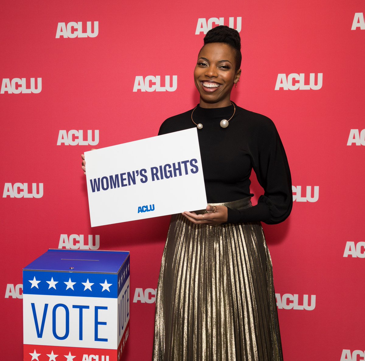 Sasheer Zamata holding a "women's rights" placard next to a ballot box