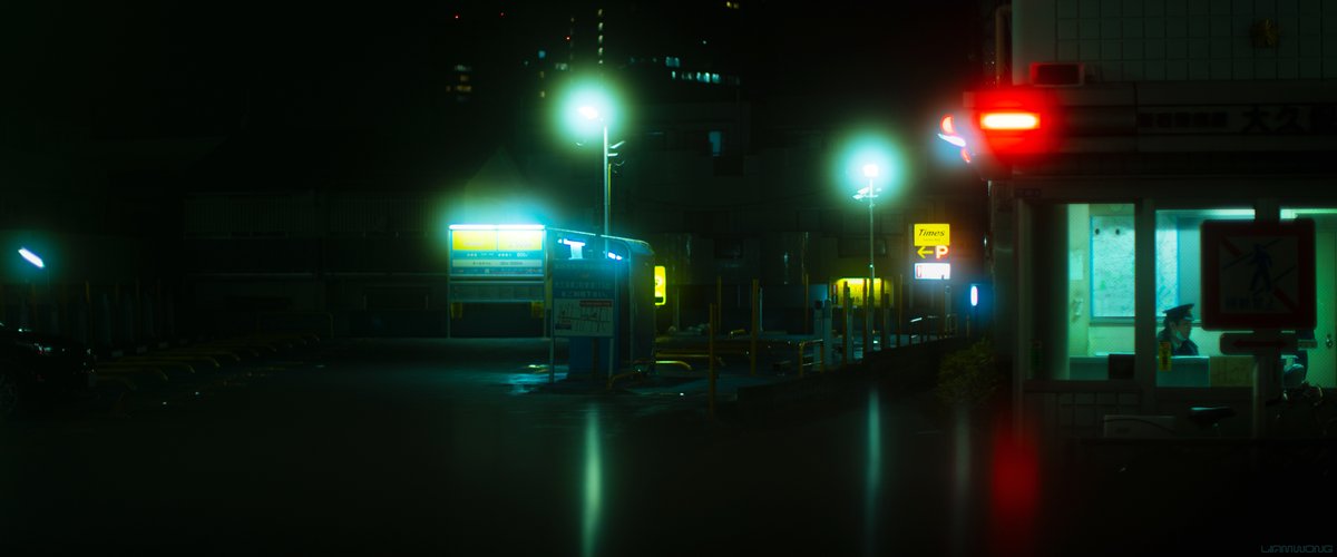 Photography by Liam Wong of Tokyo at night. A wide image of a konban place station. A cop can be seen in his office through the window. He is small in the frame to the right. The majority of the image is a car park with hazy orbs of lights caused by the filter on the lens. It feels nostalgic, almost filmic in texture. It is green and yellow.