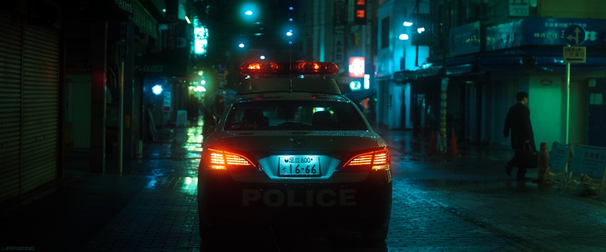 Photography by Liam Wong of Tokyo at night. A wide shot of the car in the second image but from behind. It is in the center and its lights are red. A man can be seen walking away on the right side. It is blue in color with reds and light greens.