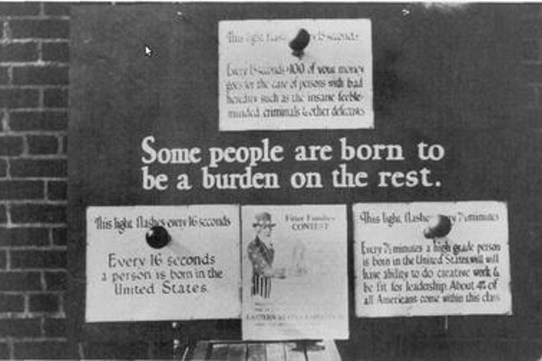 The image shows a black and white photograph of eugenics propaganda at a state fair. The poster illustrated the supposed cost of eugenically unfit classes to society and illuminated how dangerous it was with flashing lights.