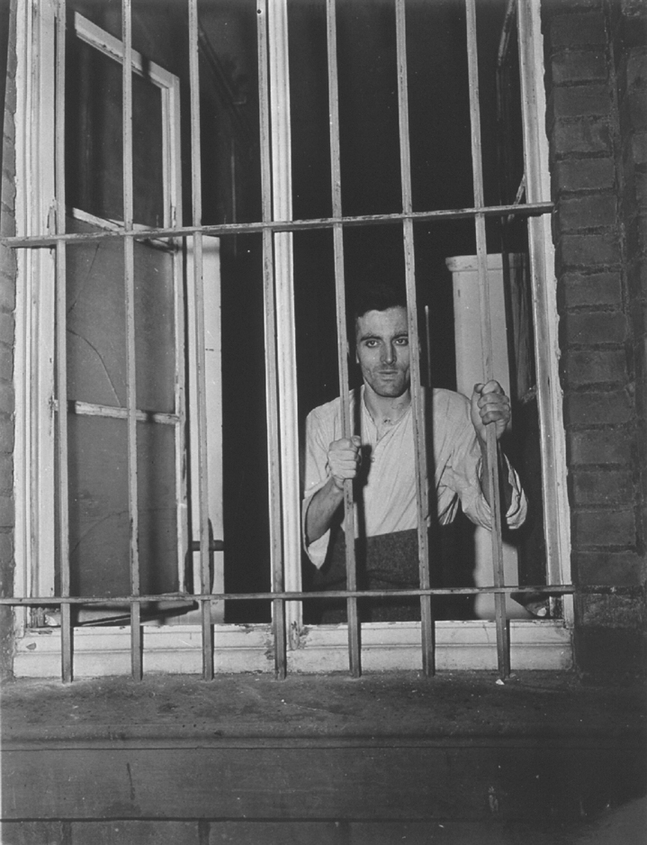 The black and white photograph shows a disabled man in a white shirt and black pants holding on to the bars of a window, at Haldemar.