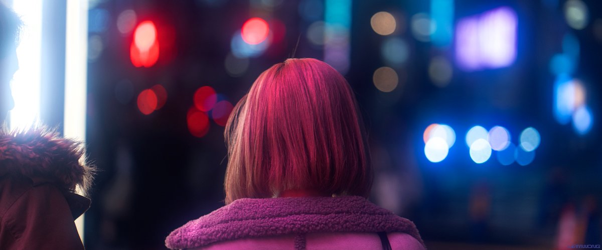 Photography by Liam Wong of Tokyo at night. An image taken standing behind a man and woman waiting to cross a street in Harajuku. The focus is on the woman, to the left the silhouette of the man speaking can be seen. He is lit up by the white light. She has a pastel pink hair color. The image is mostly blue and purple. It has a bokeh haze to it.