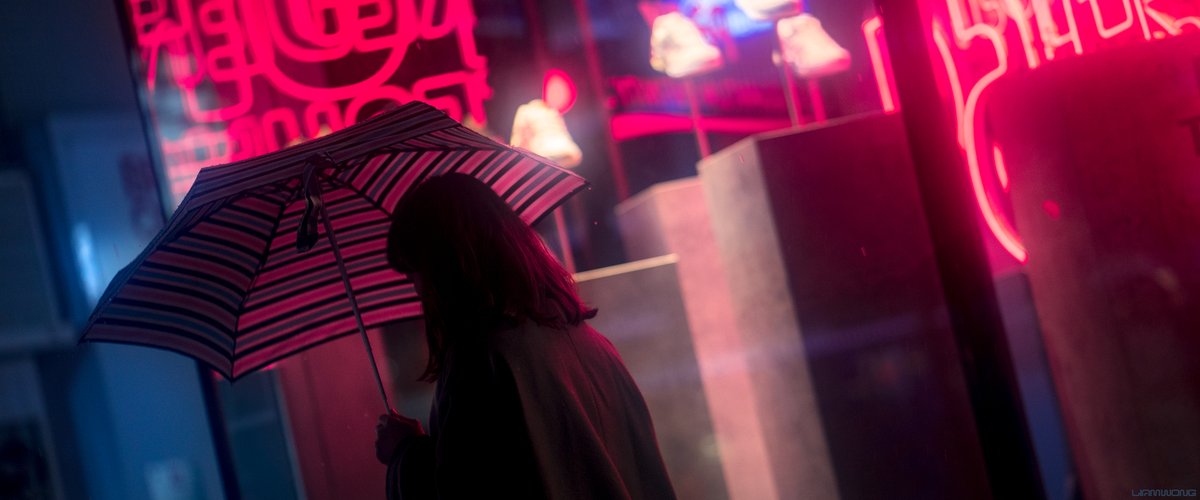 Photography by Liam Wong of Tokyo at night. A woman walking past the neon sign in the previous image. She has an umbrella which is transparent. The image is slightly off-centered which adds to the feeling that she is in a hurry to escape the rain. It is mostly pink in color.