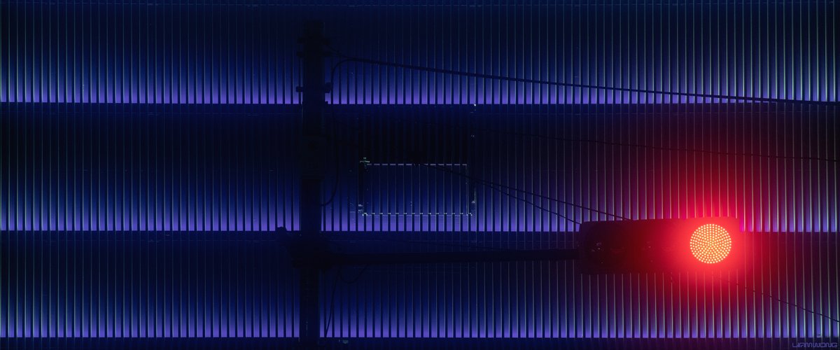 Photography by Liam Wong of Tokyo at night. An image of the front of a building in Harajuku. It feels very graphic. Many lines and grids. It is purple in color with blue tints. A traffic light adds complimentary color to the image. It has a soft red glow.